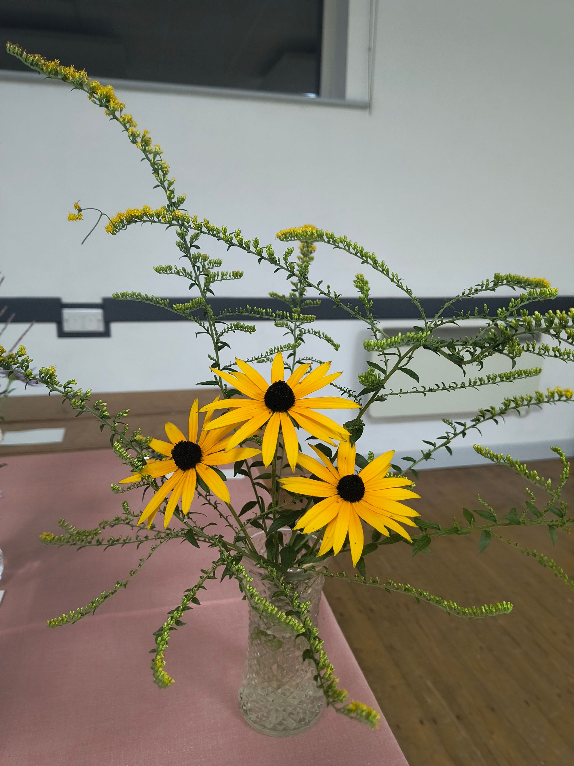Photo shows 3 orange flowers with sprigs of yellow leaves behind in a glass vase
