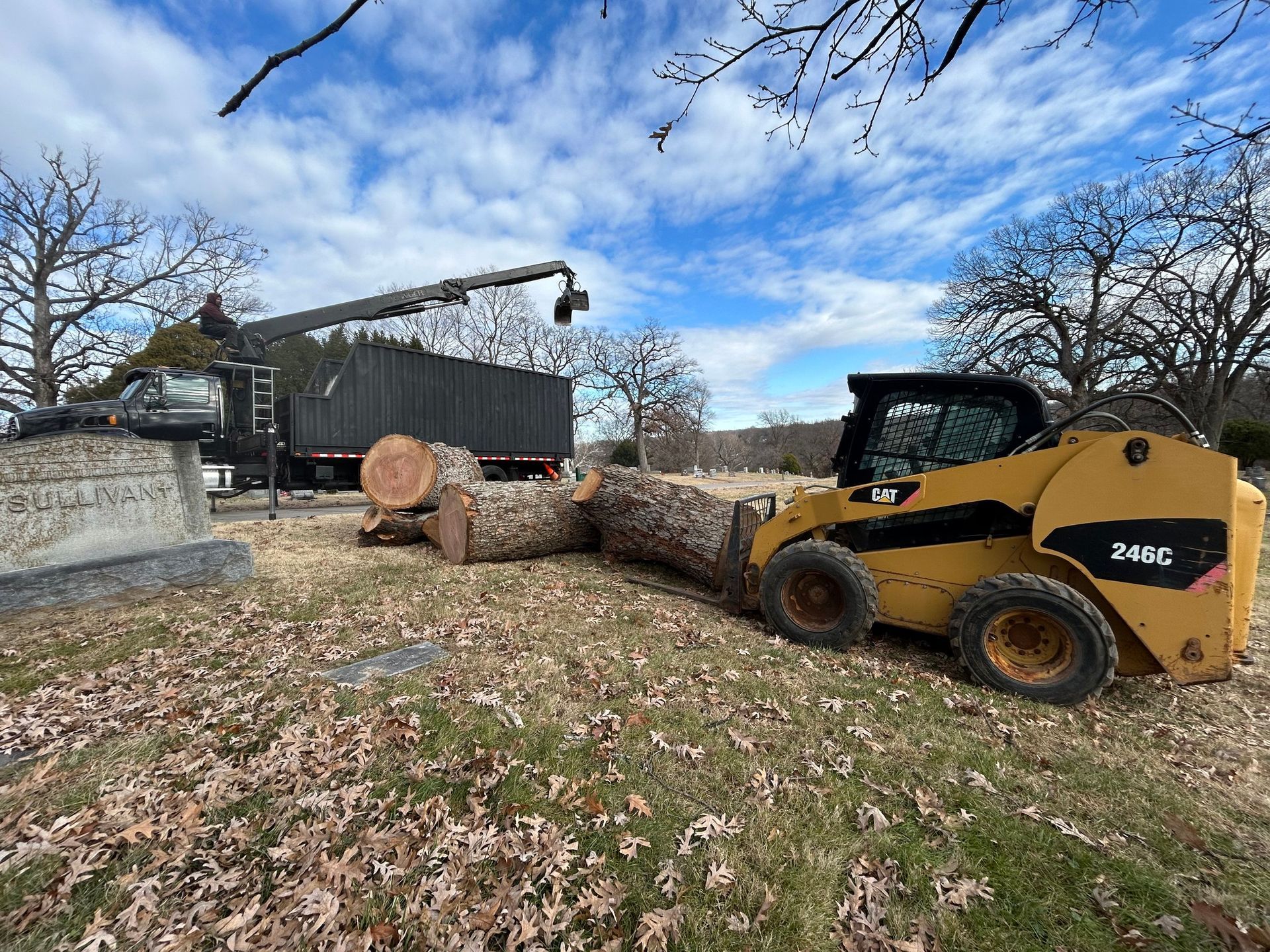 A skid steer loader moving logs near headstones with a tree-trimming truck in the background. A skid steer loader moving logs near headstones with a tree-trimming truck in the background.