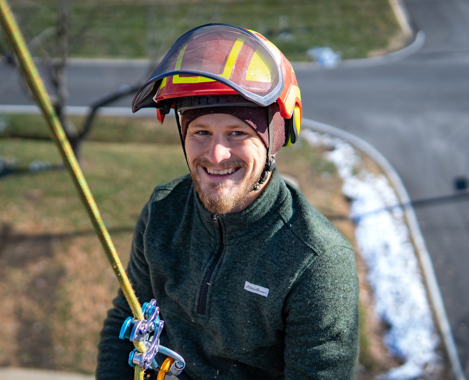 Smiling person in safety helmet and green sweater on a rope. Outdoor setting with road and snow. Smiling person in safety helmet and green sweater on a rope. Outdoor setting with road and snow.