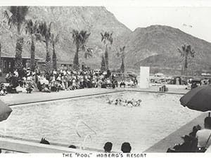 A black and white photo of a swimming pool with palm trees and mountains in the background.