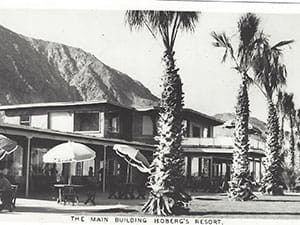 A black and white photo of a hotel with palm trees and umbrellas.