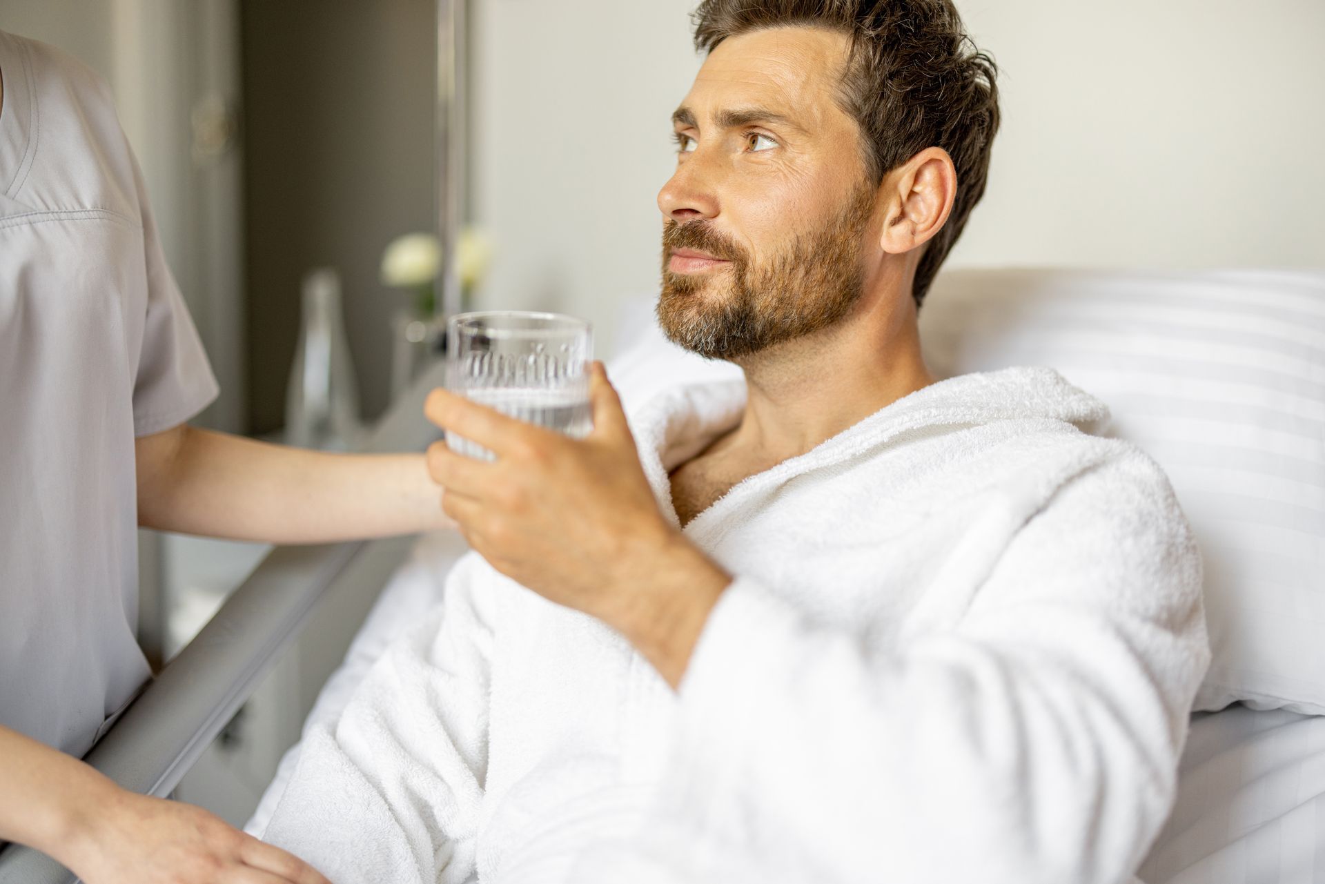 A person in a white robe holds a glass of water while sitting in bed, attended by a caregiver in a light-colored uniform.