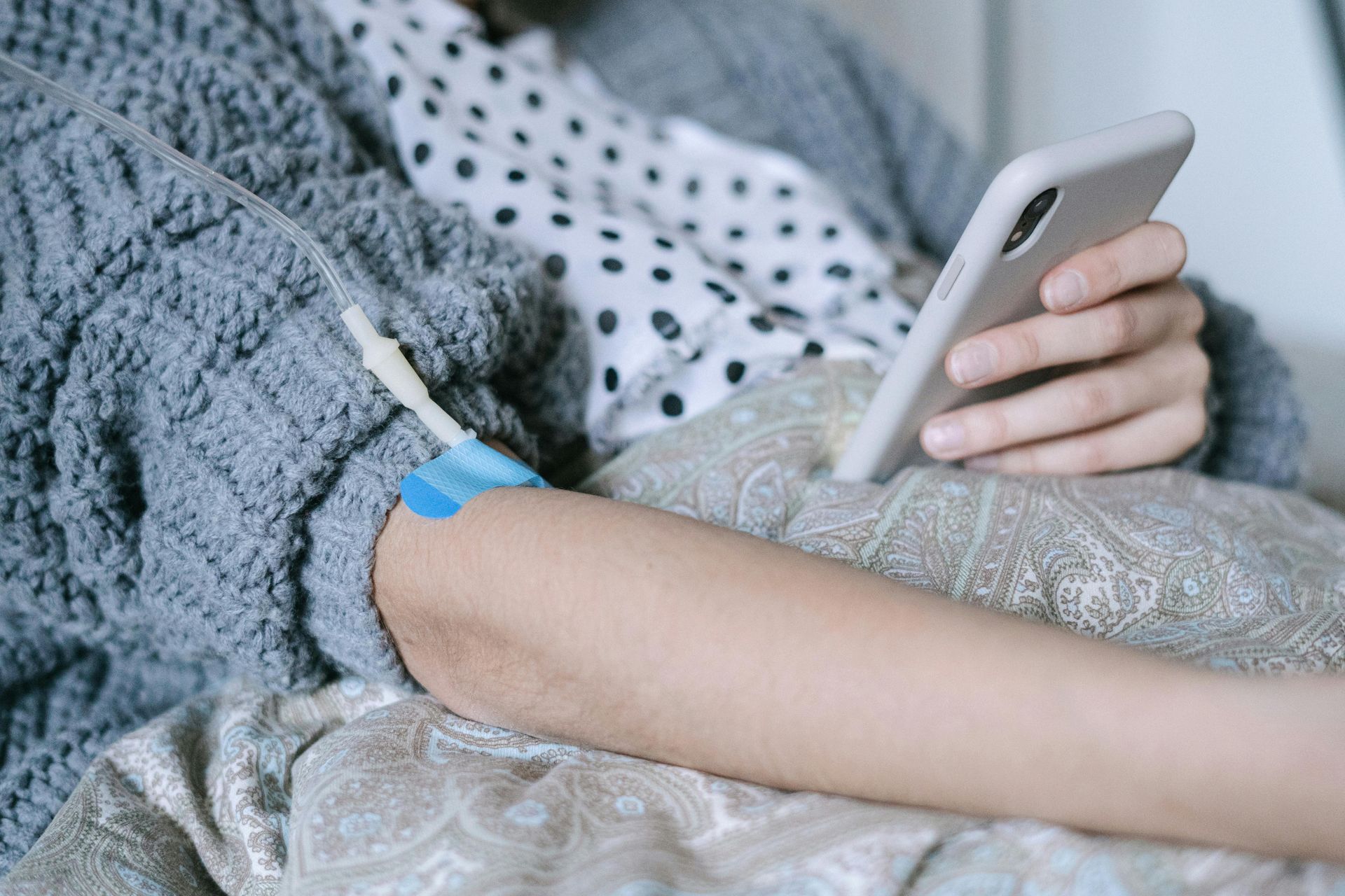 Person with an IV in arm, holding a phone, sitting on a bed or couch, wearing a sweater.