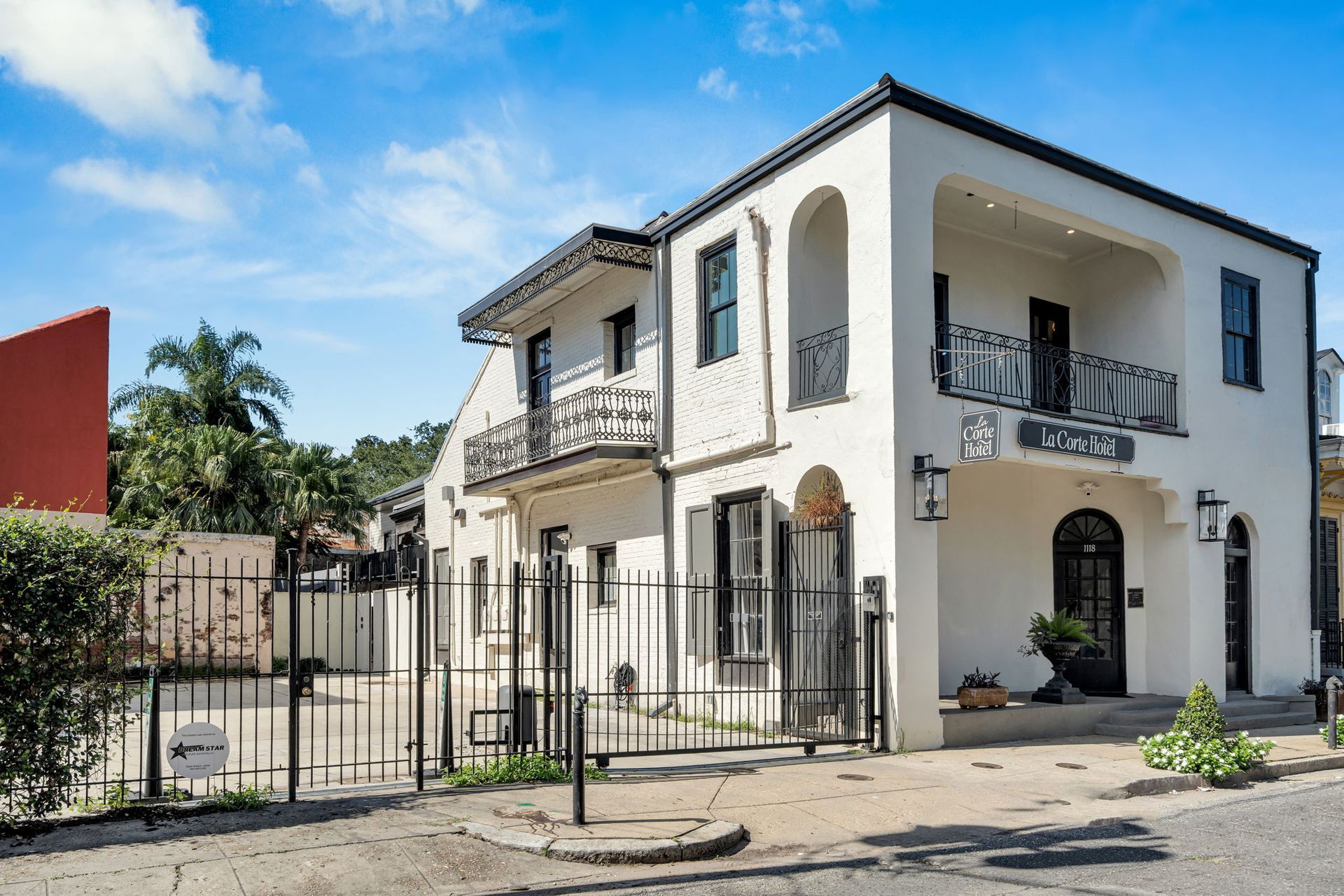 White two-story house on a sunny street corner with black balcony railings and potted plants