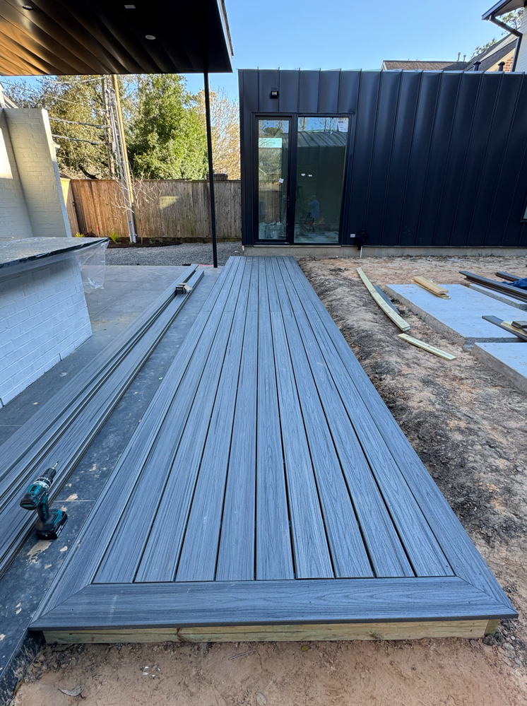 Wooden backyard deck with steps, railing, and potted plants beside a grassy yard and paved path
