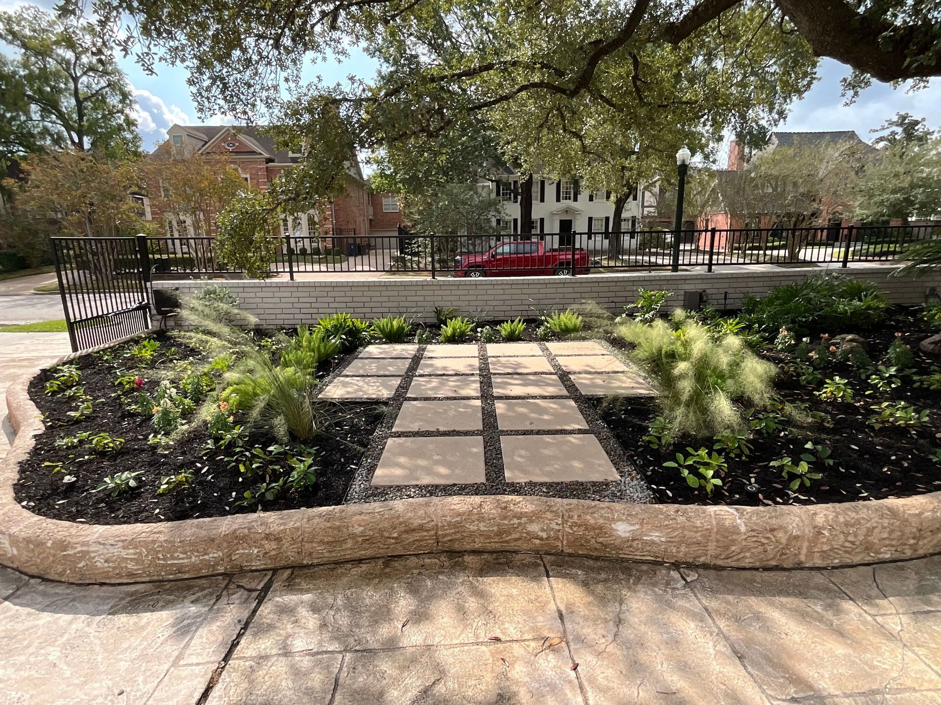Stone walkway and flower beds in a park with large tree, benches, and buildings in the background.