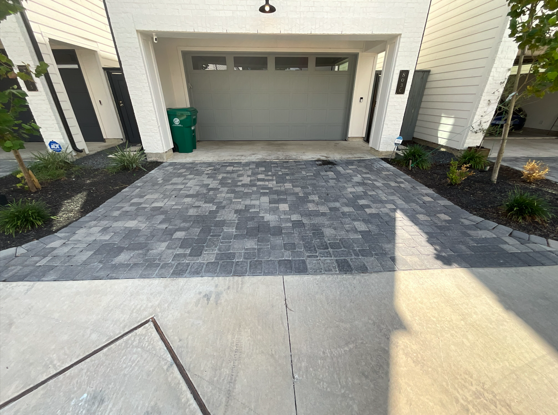 Modern house driveway with gray pavers leading to a closed garage, bordered by small landscaped beds.