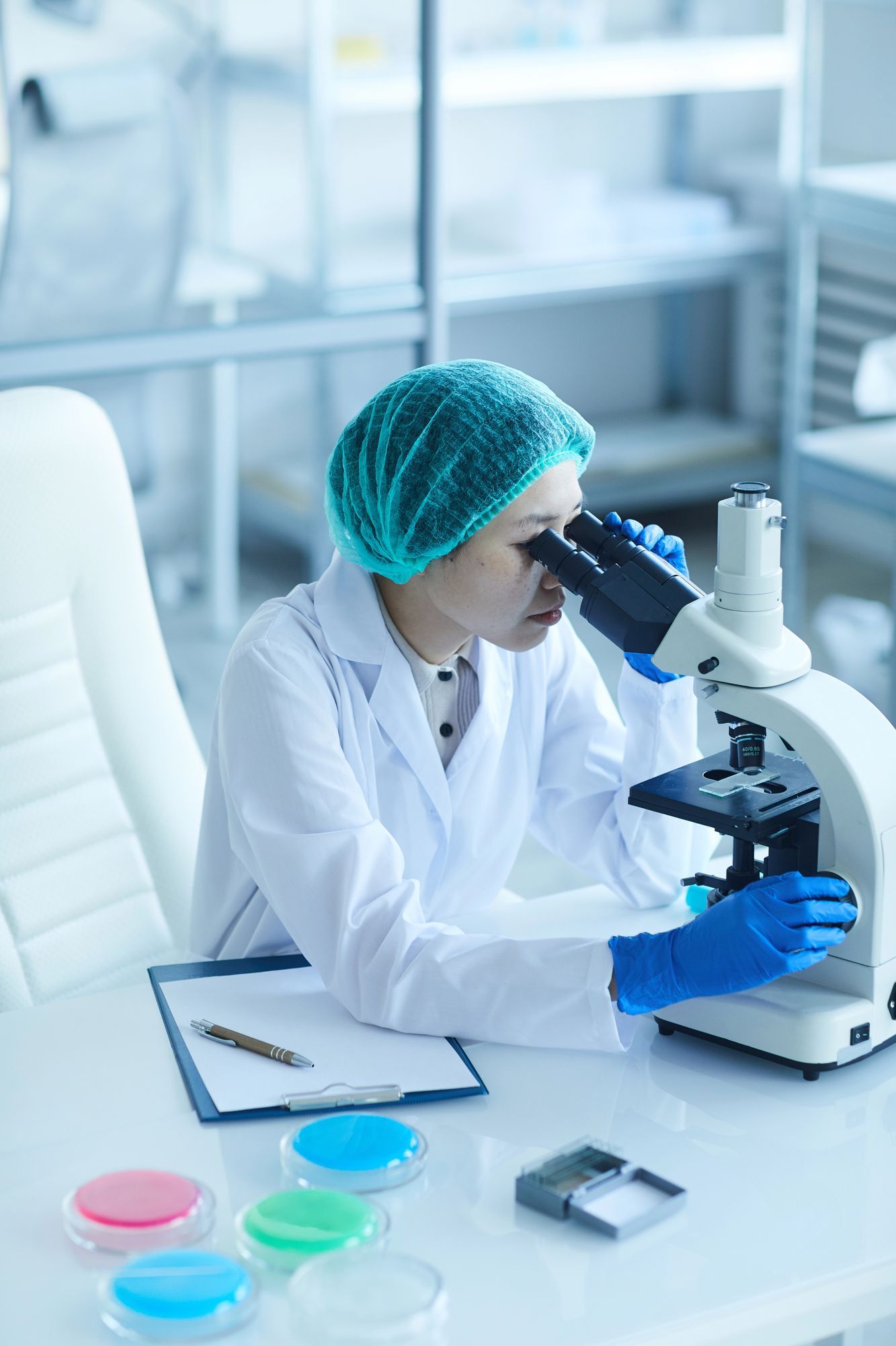 Scienziato in camice e cuffia chirurgica che esamina al microscopio campioni in piastre di Petri.