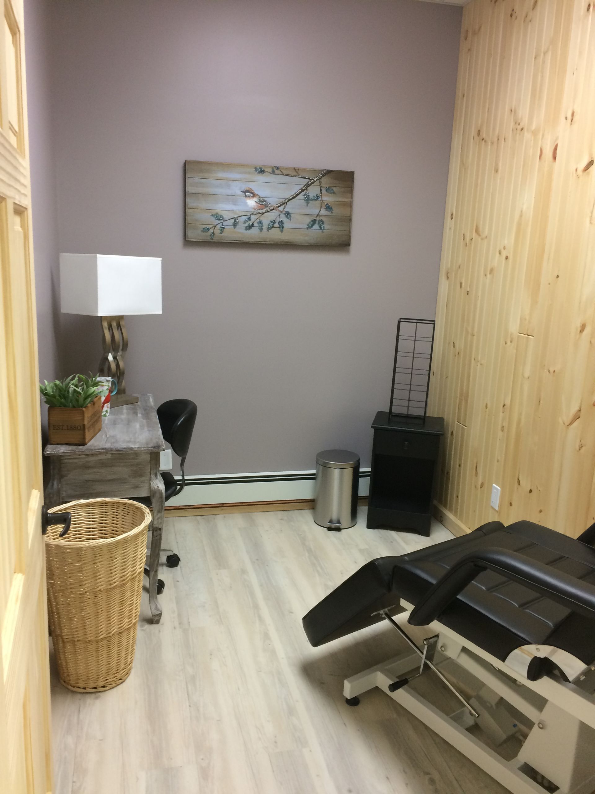 A spa treatment room featuring a reclining table, a wooden desk with a lamp, a wicker basket, and a wall art piece.