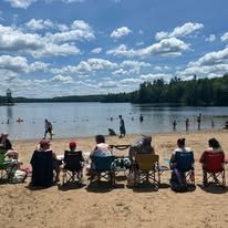 People in camping chairs sit on a sandy beach looking out at a lake with swimmers under a bright, cloudy blue sky.
