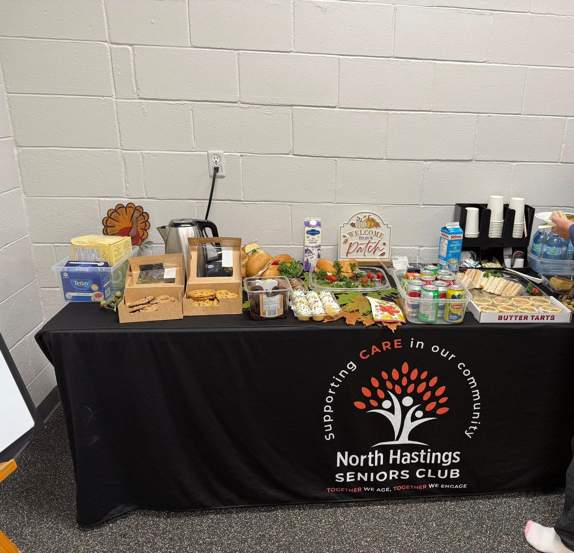 A table covered in a black North Hastings Seniors Club cloth, set with food, drinks, and snacks against a white wall.