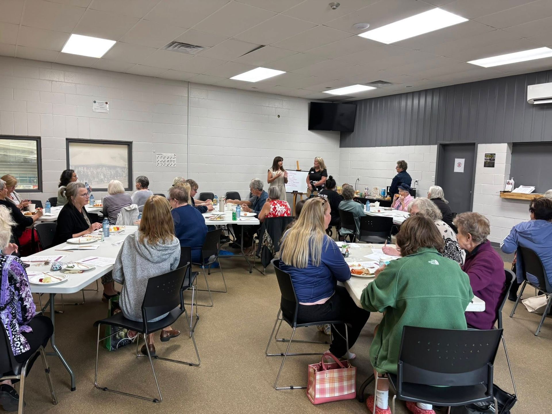 A group of people sit at several tables in a community room, attending a presentation or workshop.