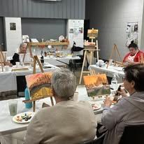 A painting class in a bright room where people sit at tables with canvases on easels, working on sunset landscapes.