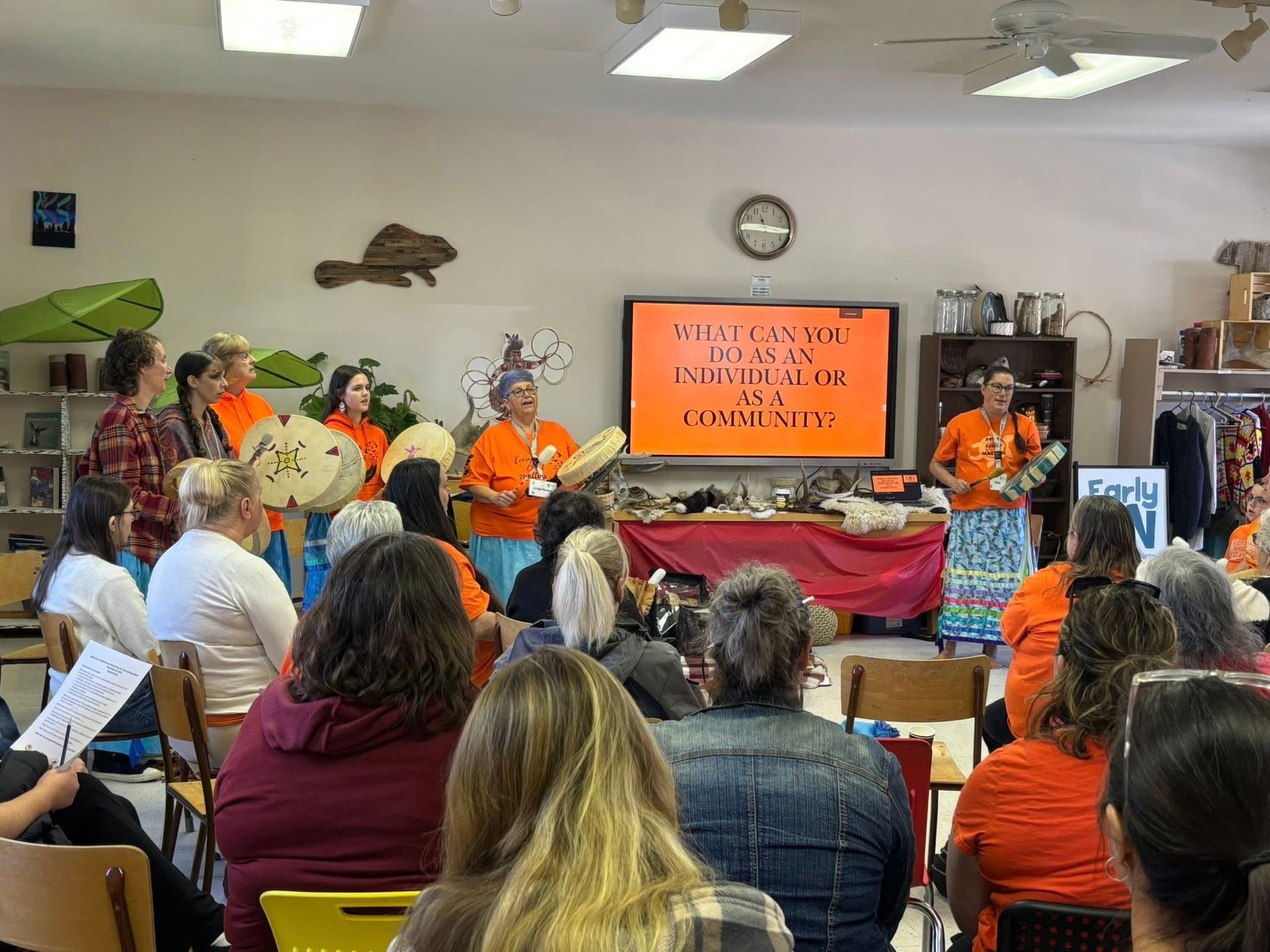 A group of people wearing orange shirts gather in a classroom for a presentation with a screen displaying text.