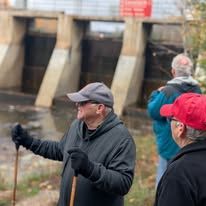 Three people stand near a water dam, with one person in the foreground gesturing toward it with a walking stick.