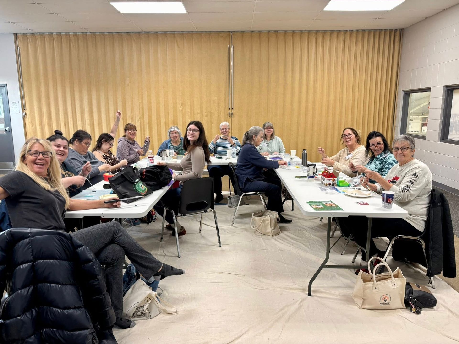 A diverse group of people sitting at tables in a brightly lit room, smiling and crafting together.
