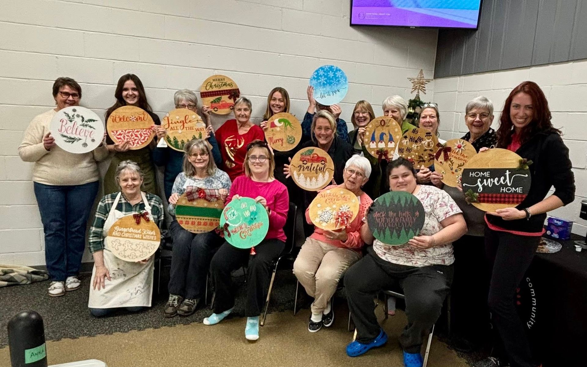 A group of people standing and sitting in a room, holding up their hand-painted, circular wooden signs with festive designs.
