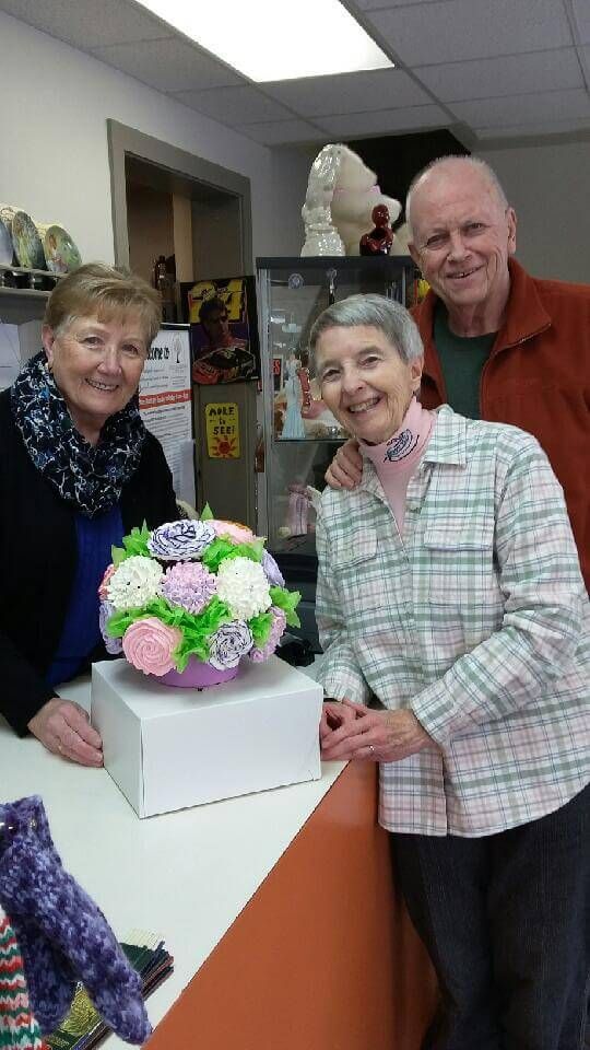 Three people smile behind a white counter with a colorful cupcake bouquet centerpiece in a brightly lit room.