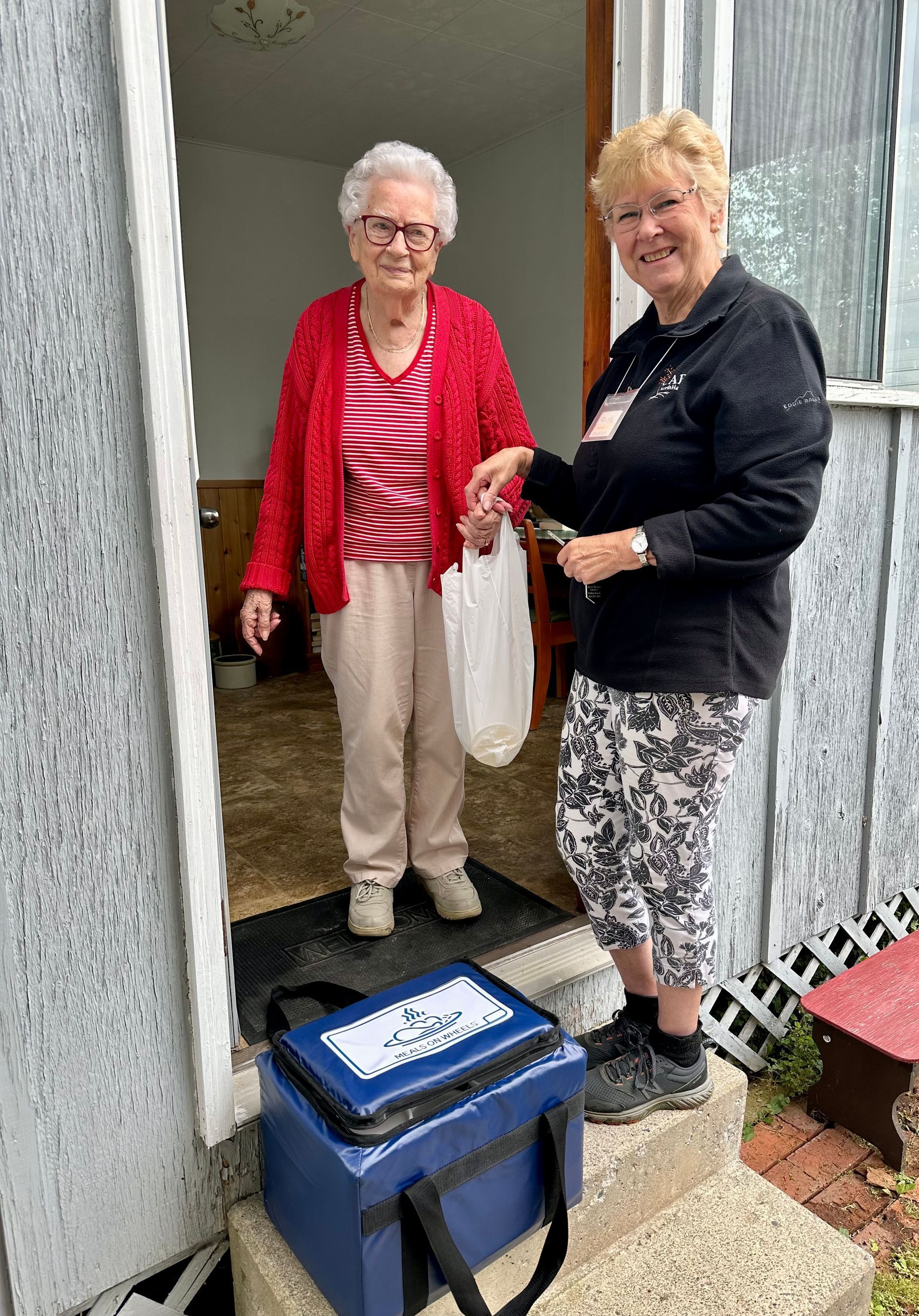 A volunteer hands a white bag to a resident at their doorway, with a blue delivery cooler sitting on the porch steps.