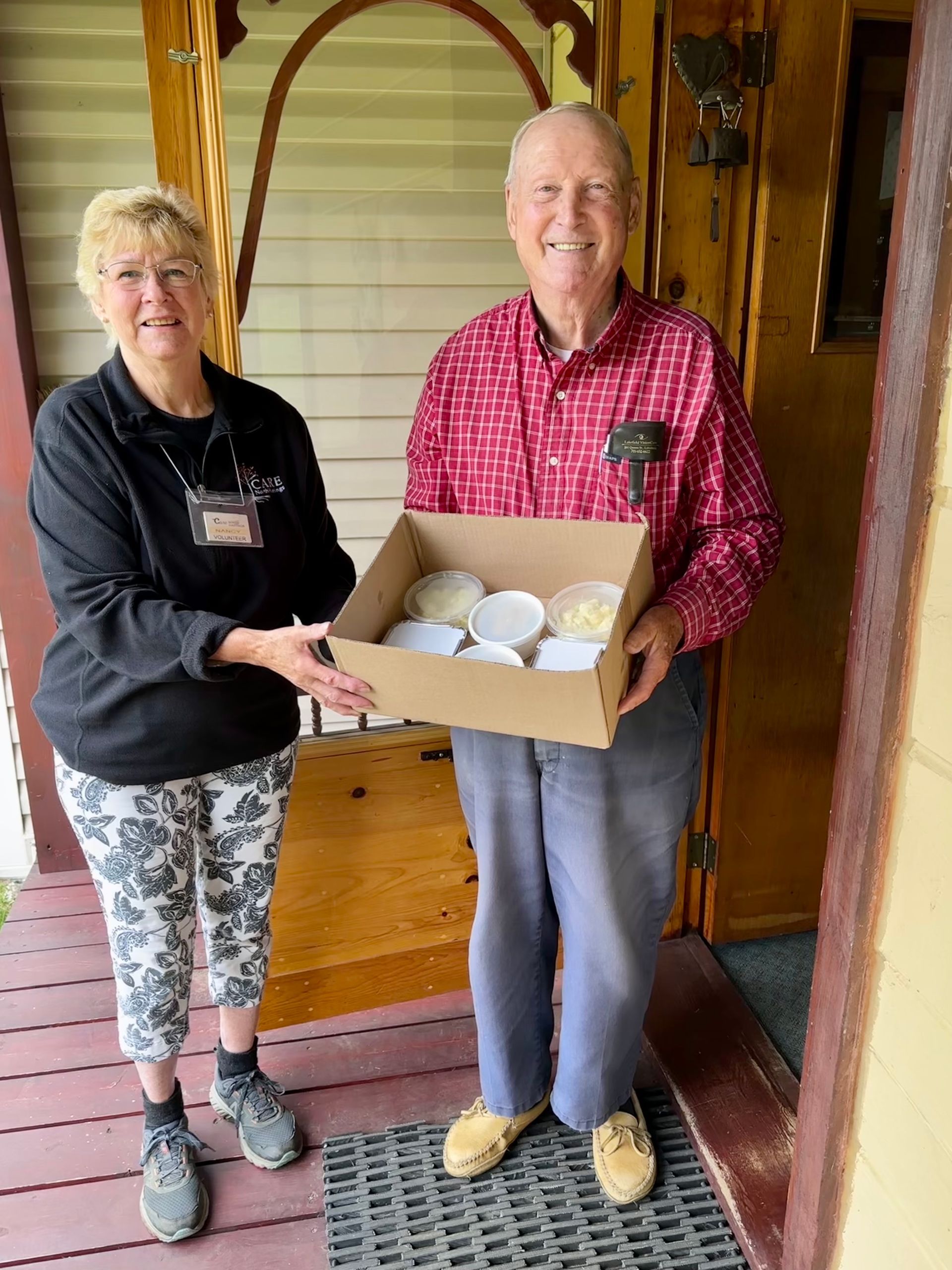 Two smiling people hold a cardboard box of food containers on a front porch.