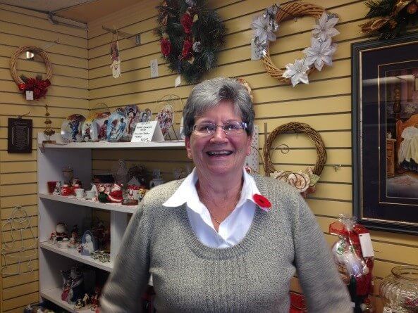 A smiling person stands in a gift shop in front of wall-mounted wreaths and shelves filled with holiday decorations.