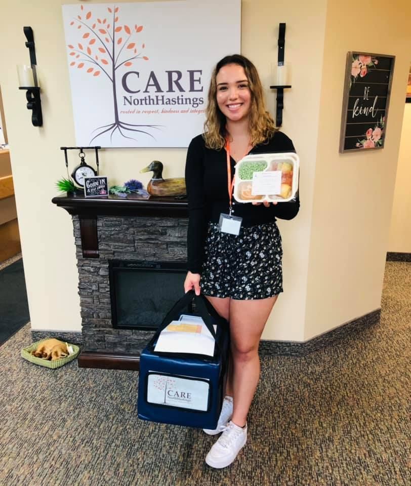 A smiling person holds a meal container and a CARE North Hastings branded delivery bag in front of a fireplace and logo.