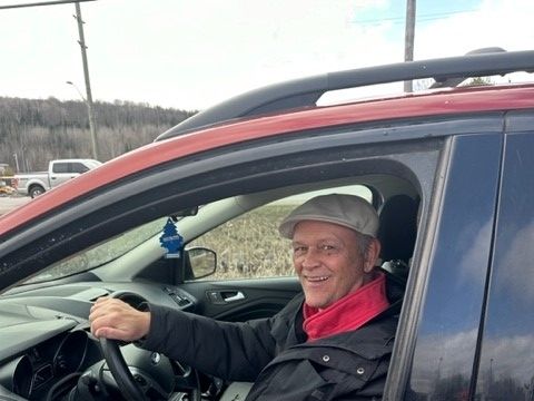 A smiling person wearing a cap and jacket driving a red car with a hanging air freshener, seen from the passenger side.