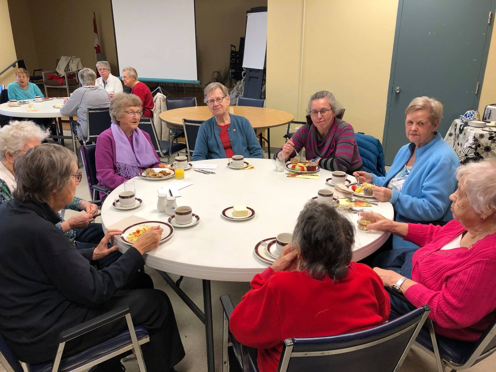 A group of people sit around a large, circular table in a community room, eating a meal and conversing.
