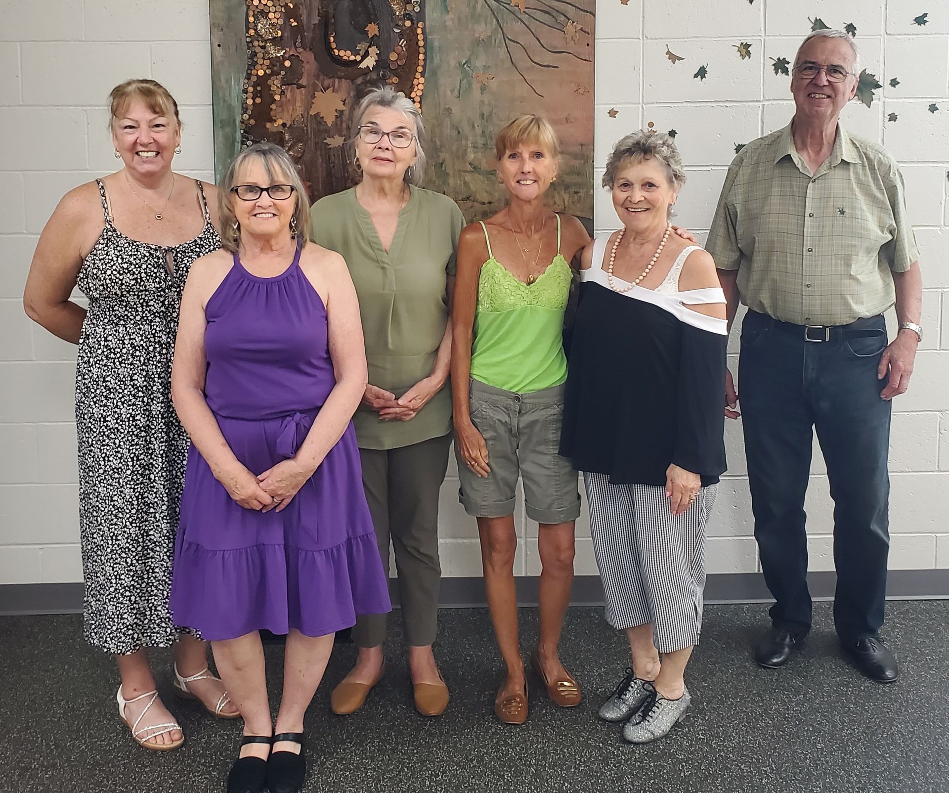 A group of six people stand side-by-side smiling in front of a white tiled wall with a vertical art piece.