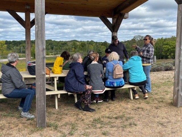 A group sits at a picnic table under a wooden pavilion while a person stands and speaks to them in an outdoor field.