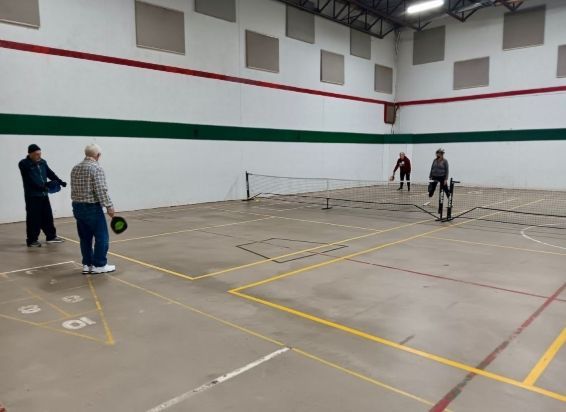 Four people play pickleball on an indoor court with white walls and green horizontal stripes.