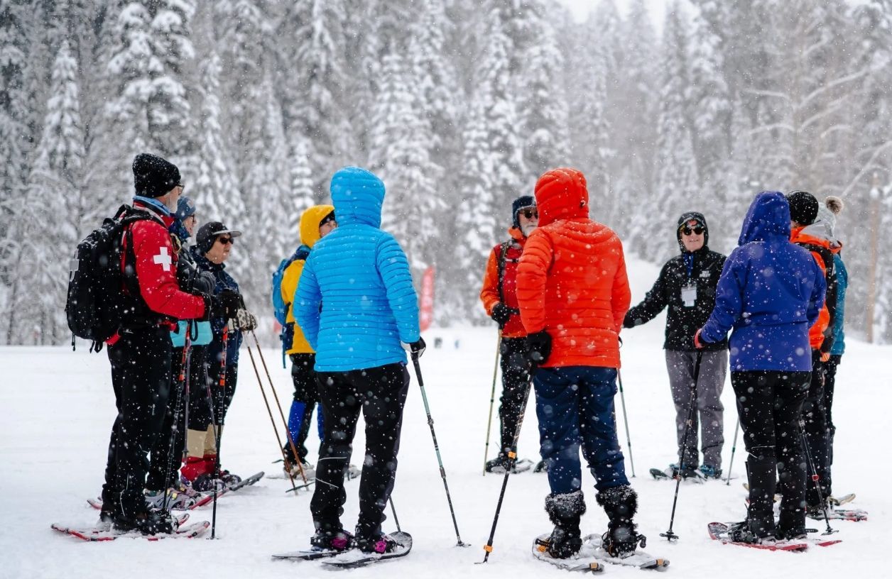 A group of people in colorful winter gear and snowshoes gather in a snowy forest for a guided activity.