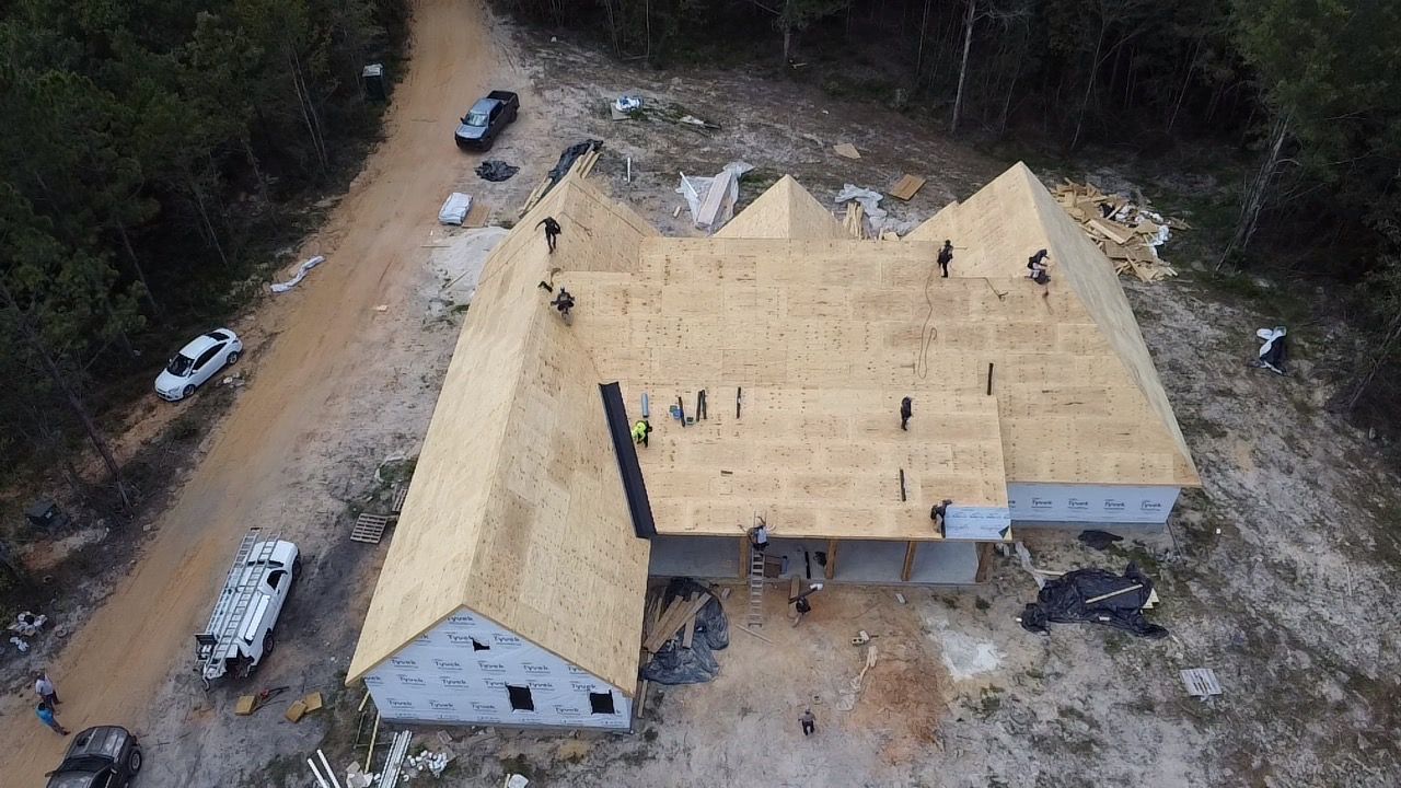 Aerial view of a large house under construction with a wood roof, surrounded by dirt and trees.