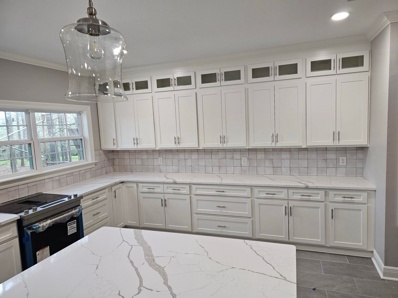 Bright white kitchen with marble island, shaker cabinets, pendant light, and large window