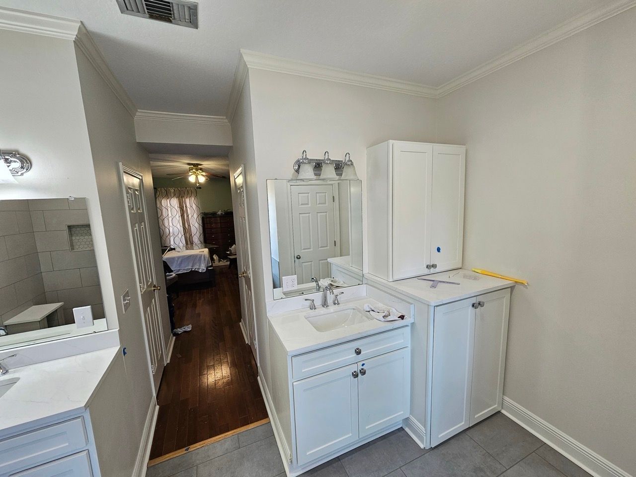 Bright bathroom with white vanity, sink, mirror, and cabinets; hallway visible beyond.