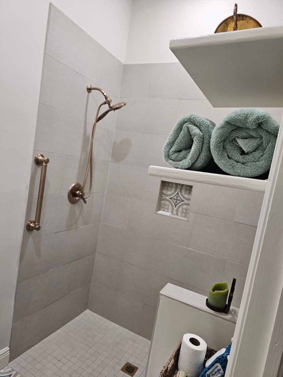 Small white bathroom shower with brass fixtures, gray tile, built-in shelves, and rolled teal towels.