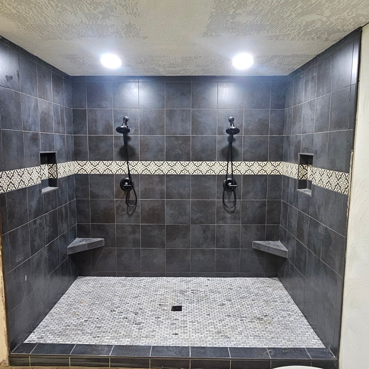 Stone-tiled walk-in shower with two showerheads, mosaic accent strip, and built-in bench under bright lights