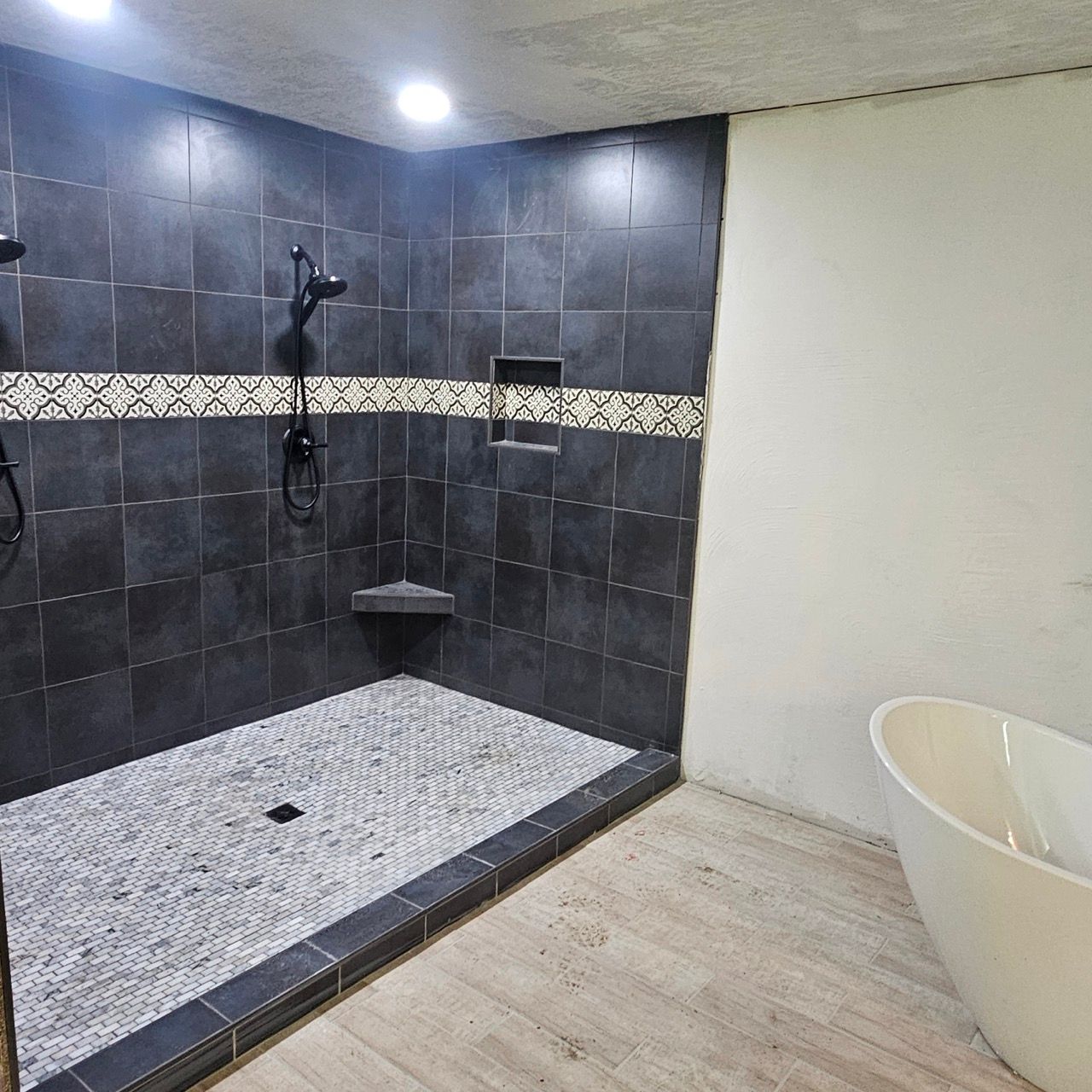 Modern shower with dark tile, mosaic floor, and a white freestanding tub beside it