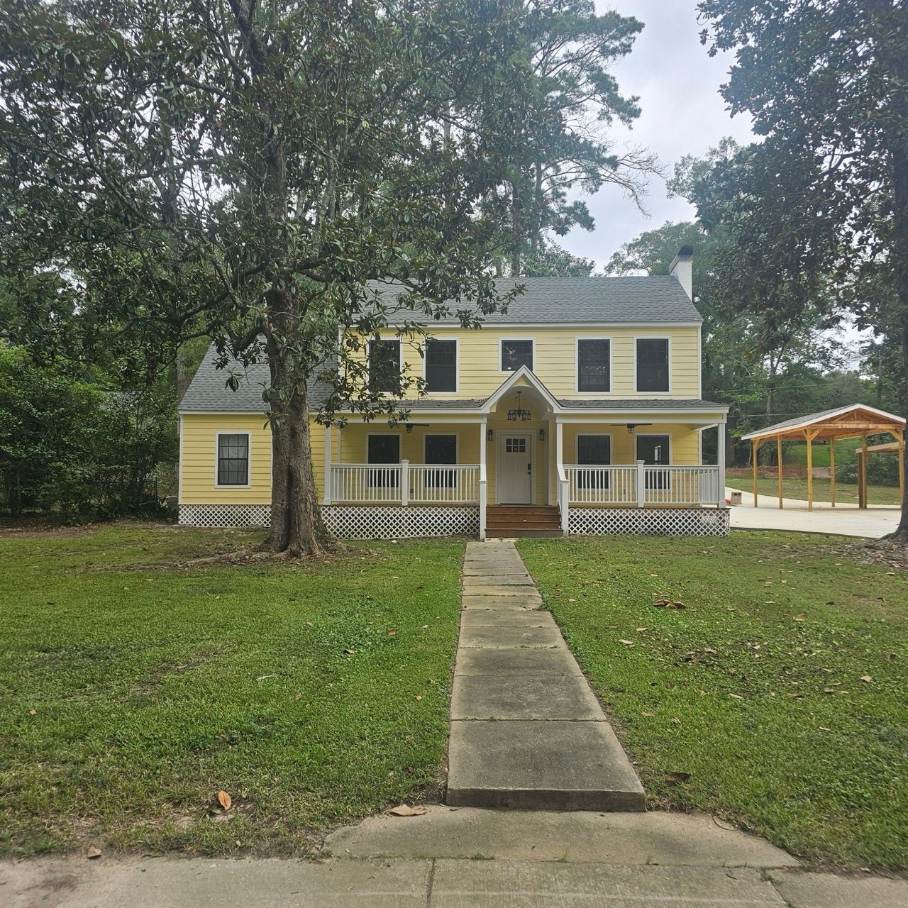 Yellow two-story house with a front porch, large trees, and a walkway leading from the street.