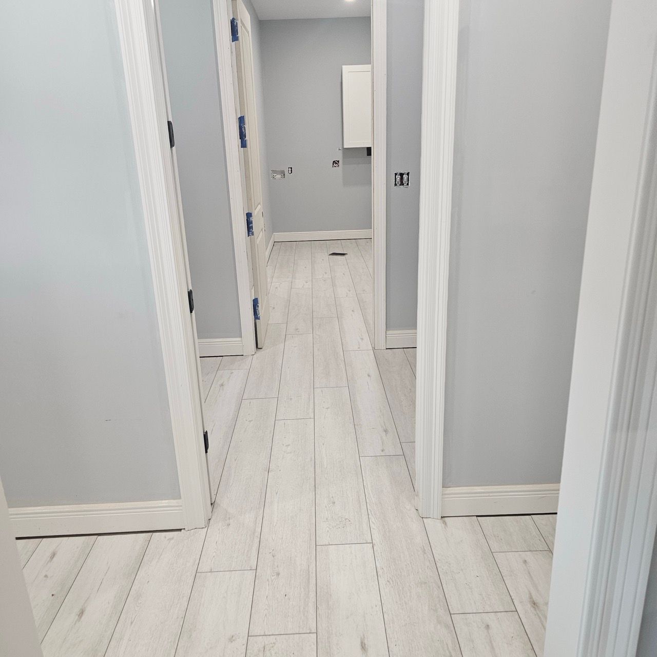 Narrow white hallway with light gray walls and wood flooring, leading to a doorway and cabinet in the distance