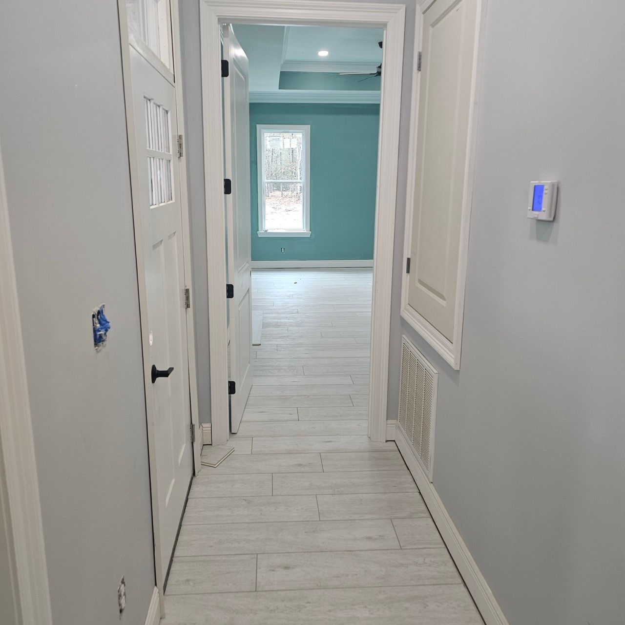 Narrow hallway with white walls and light wood floors leading to a room with a teal accent wall and window.