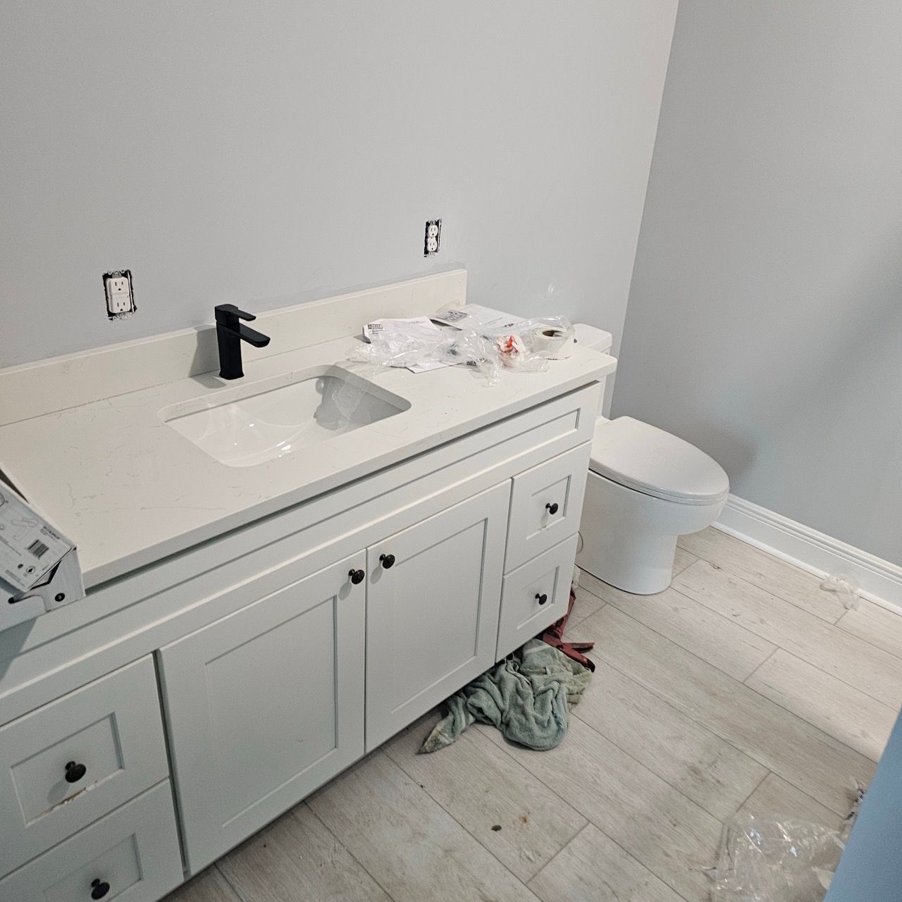 Bathroom vanity with sink, black faucet, white cabinets, toilet, and a trash bag on the floor.