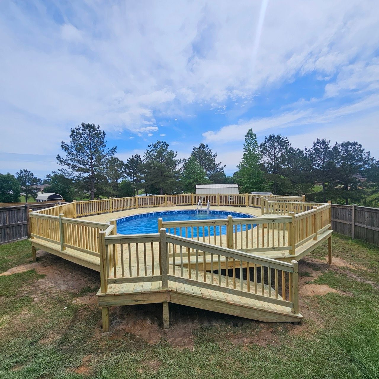 Above-ground wooden pool deck under construction in a grassy yard, with blue pool and cloudy sky.