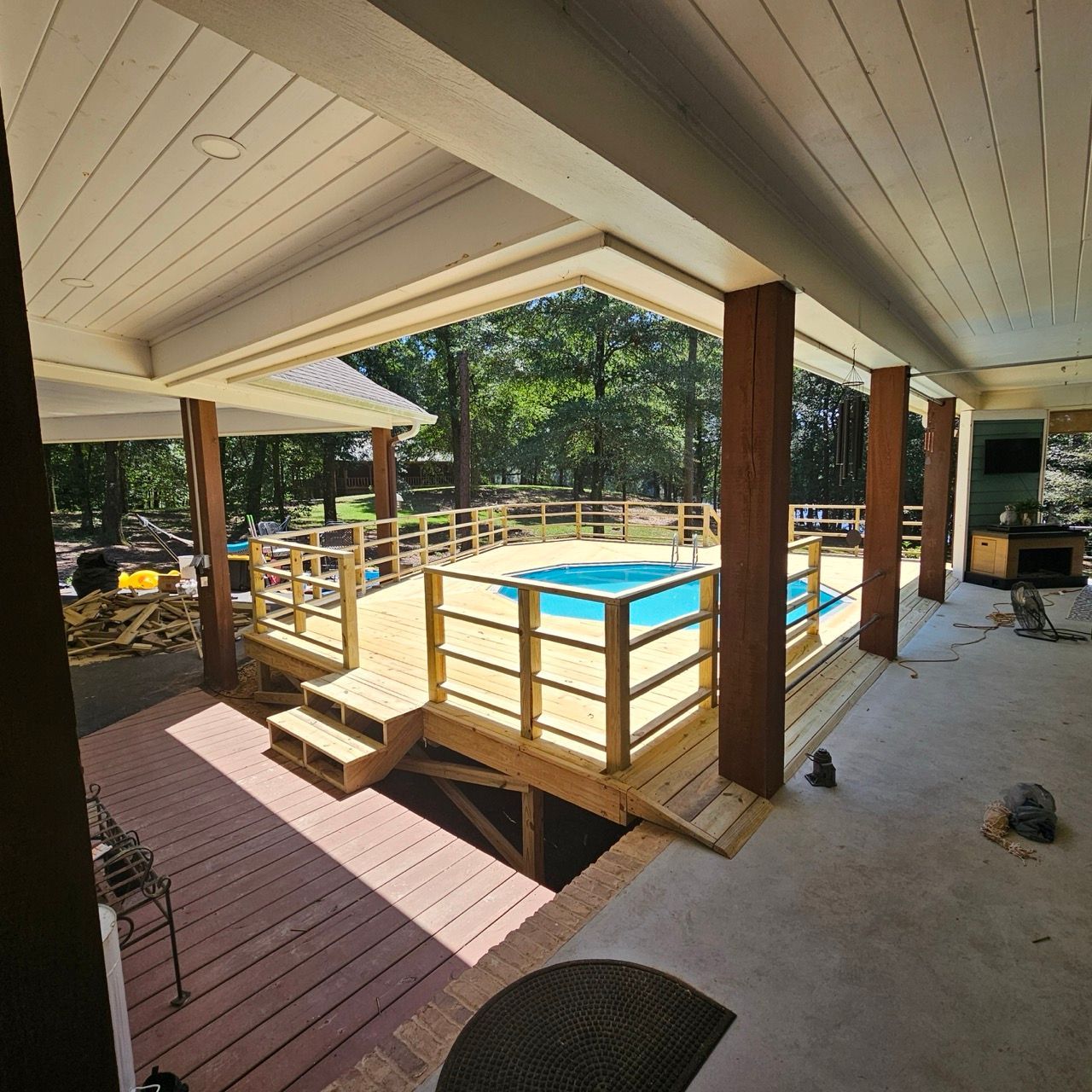 Covered backyard pool with wooden deck, railing, and steps under construction, surrounded by trees.