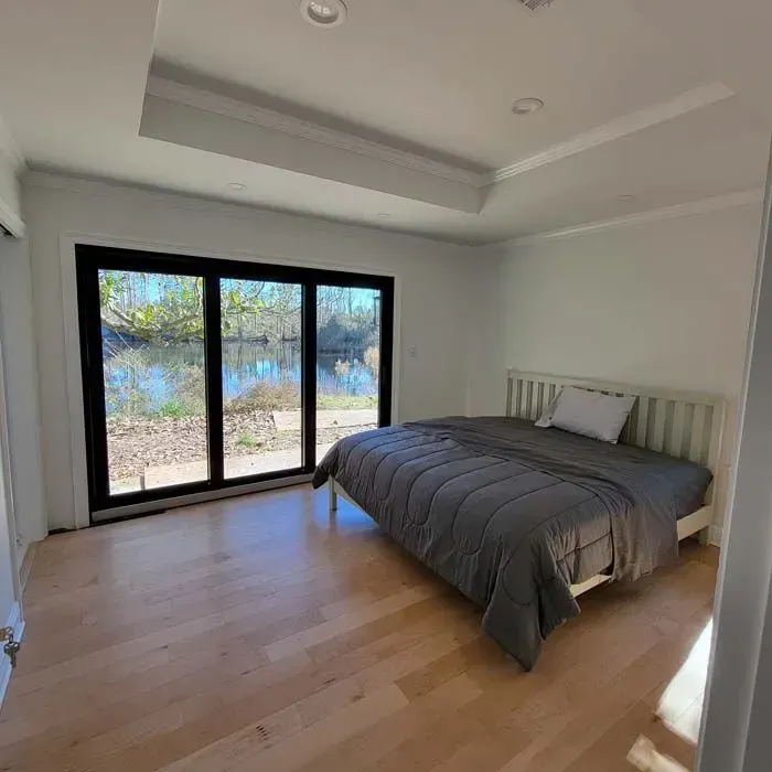 Bedroom with a white bed, gray bedding, and large black-framed glass doors overlooking a body of water.