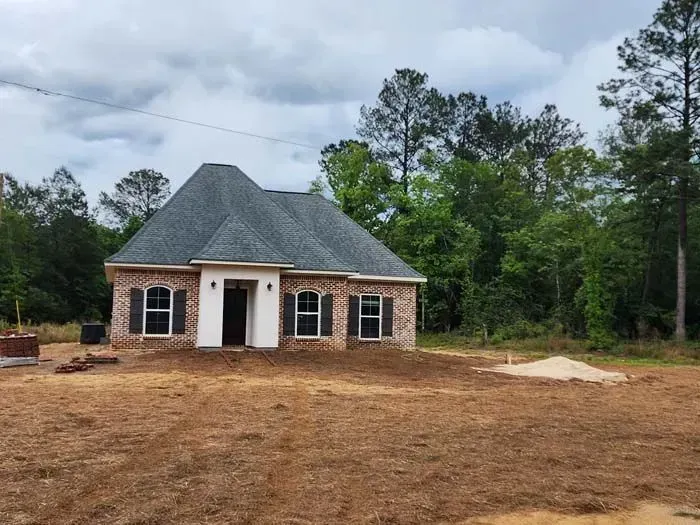 Brick house under construction with gray roof, white trim, and black shutters; set in a clearing.