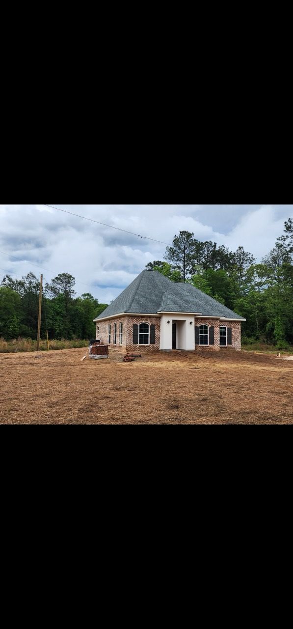 Small unfinished house with a gray roof in a cleared dirt lot, surrounded by trees under a cloudy sky