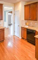 A kitchen with wooden cabinets, granite countertops, hardwood floors, and a black dishwasher, with an open doorway.