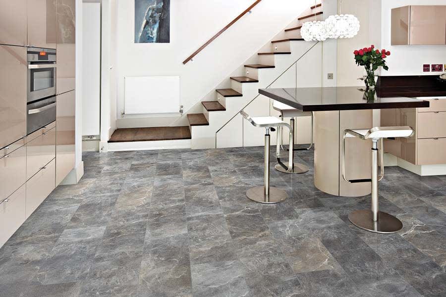 A modern kitchen with grey slate-look tile flooring, a dining peninsula with two white bar stools, and a wooden staircase.