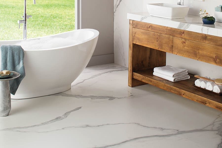 Modern bathroom with a white freestanding tub, marble-patterned tile floor, and a wooden vanity with white towels.